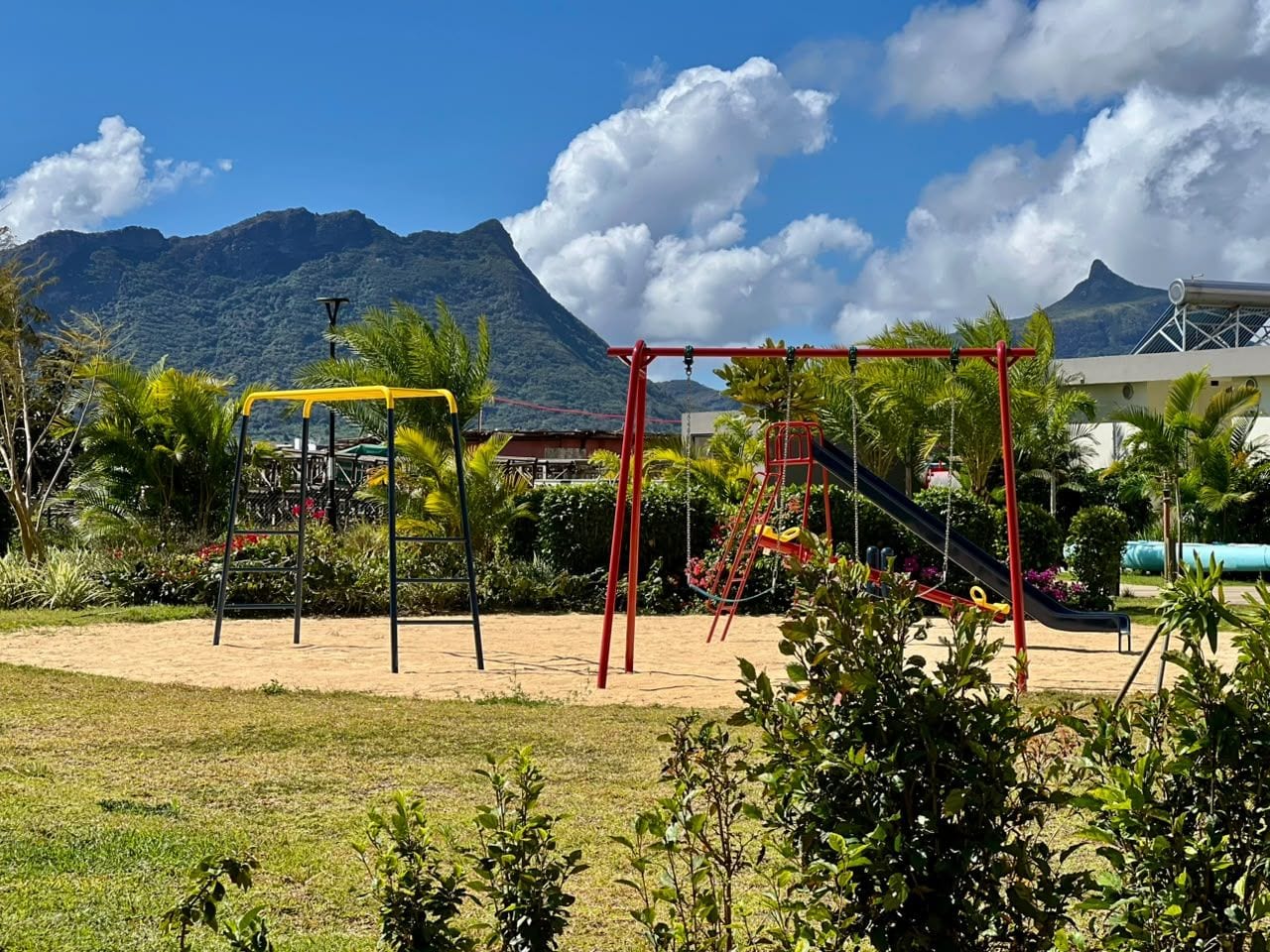 A playground with swings and slides, surrounded by greenery and a mountain in the background.