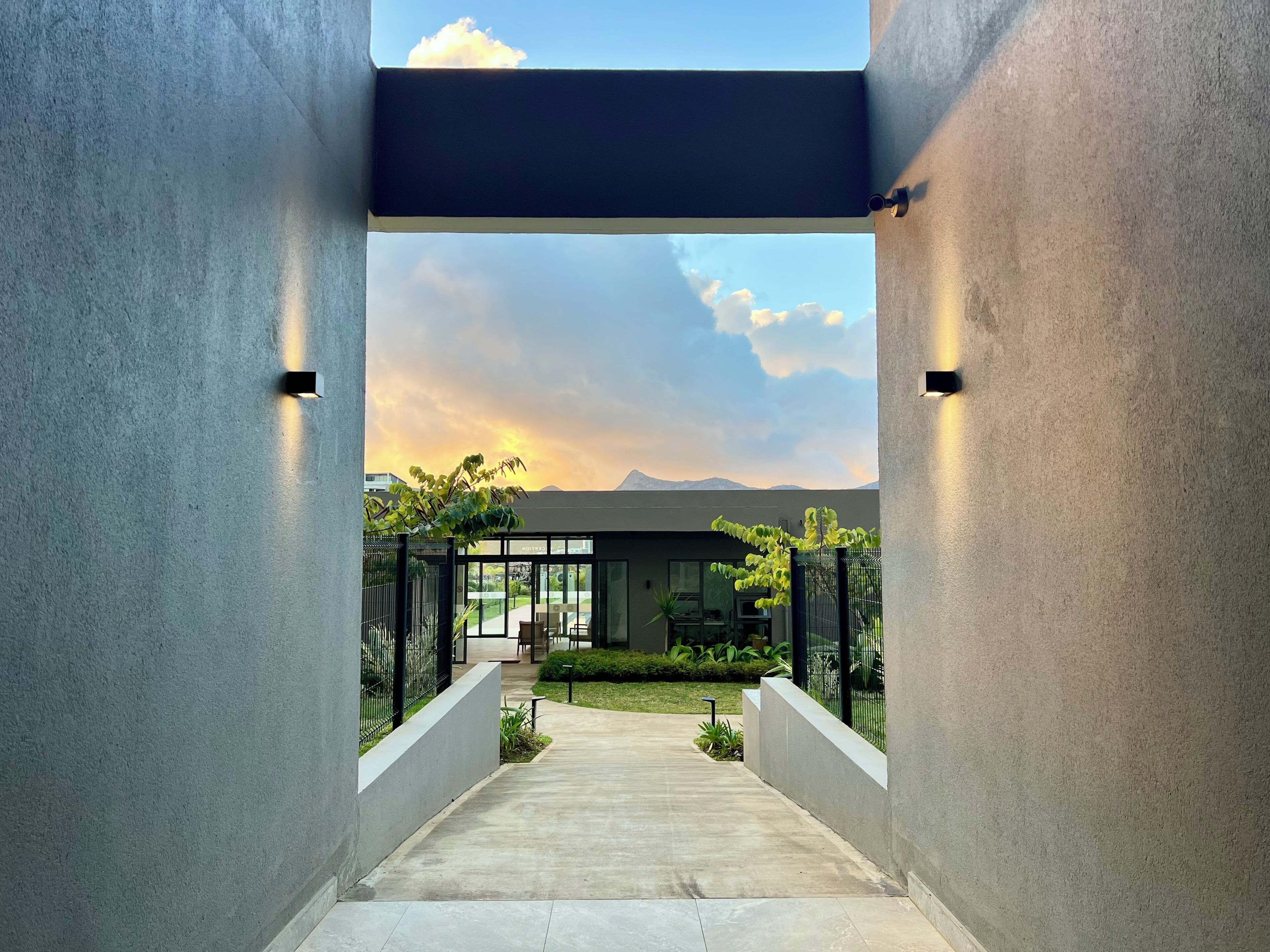 Entrada moderna de un edificio con un sendero de concreto que lleva a una puerta de vidrio, rodeada de follaje y un cielo despejado.