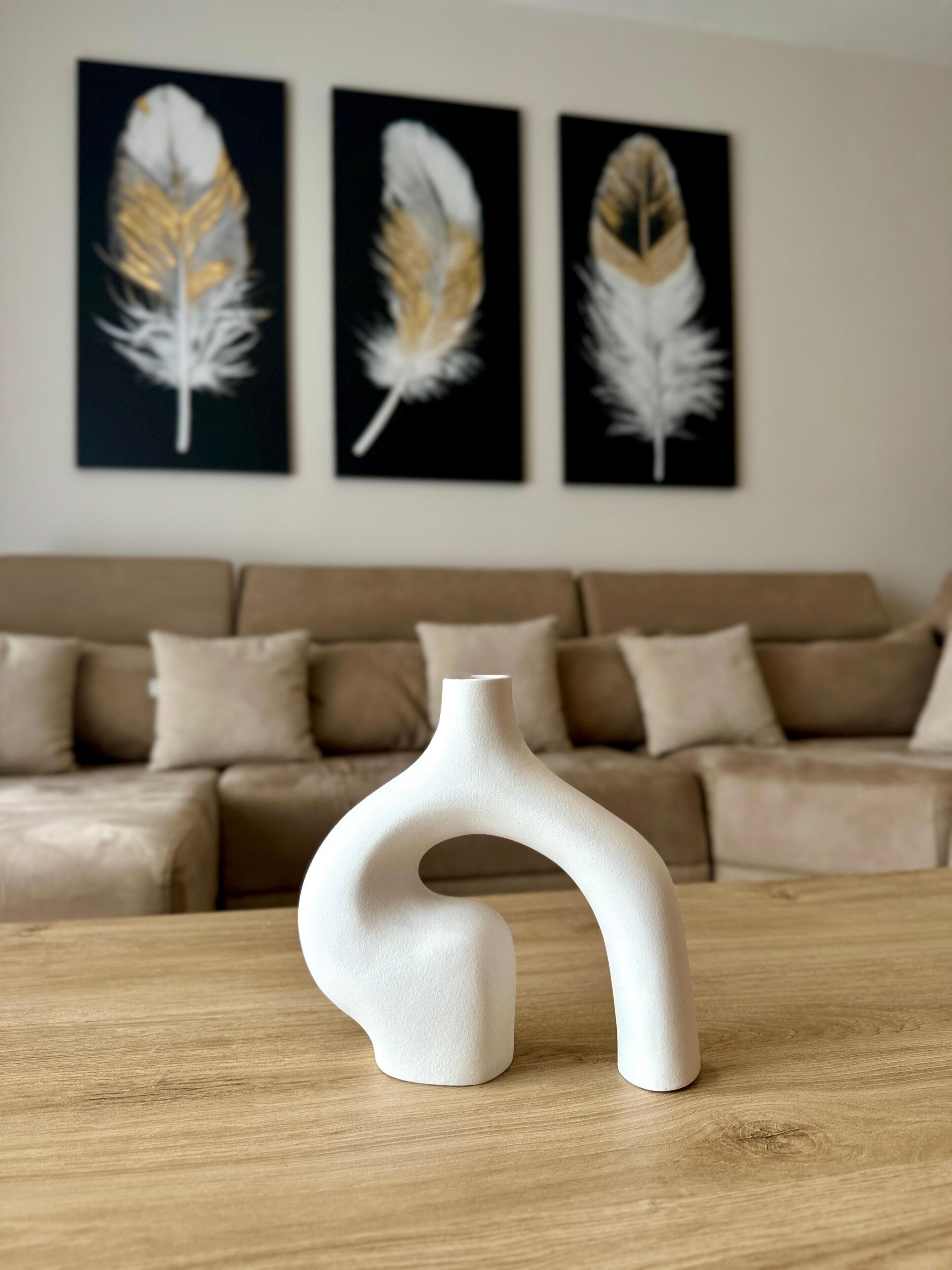 Een moderne woonkamer met een witte vaas op een houten tafel, een beige bank en drie decoratieve veren aan de wand.
