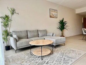A modern living room with a grey sectional sofa, two round wooden coffee tables, a large abstract painting, and potted plants. - Minissy Suites (Moka)