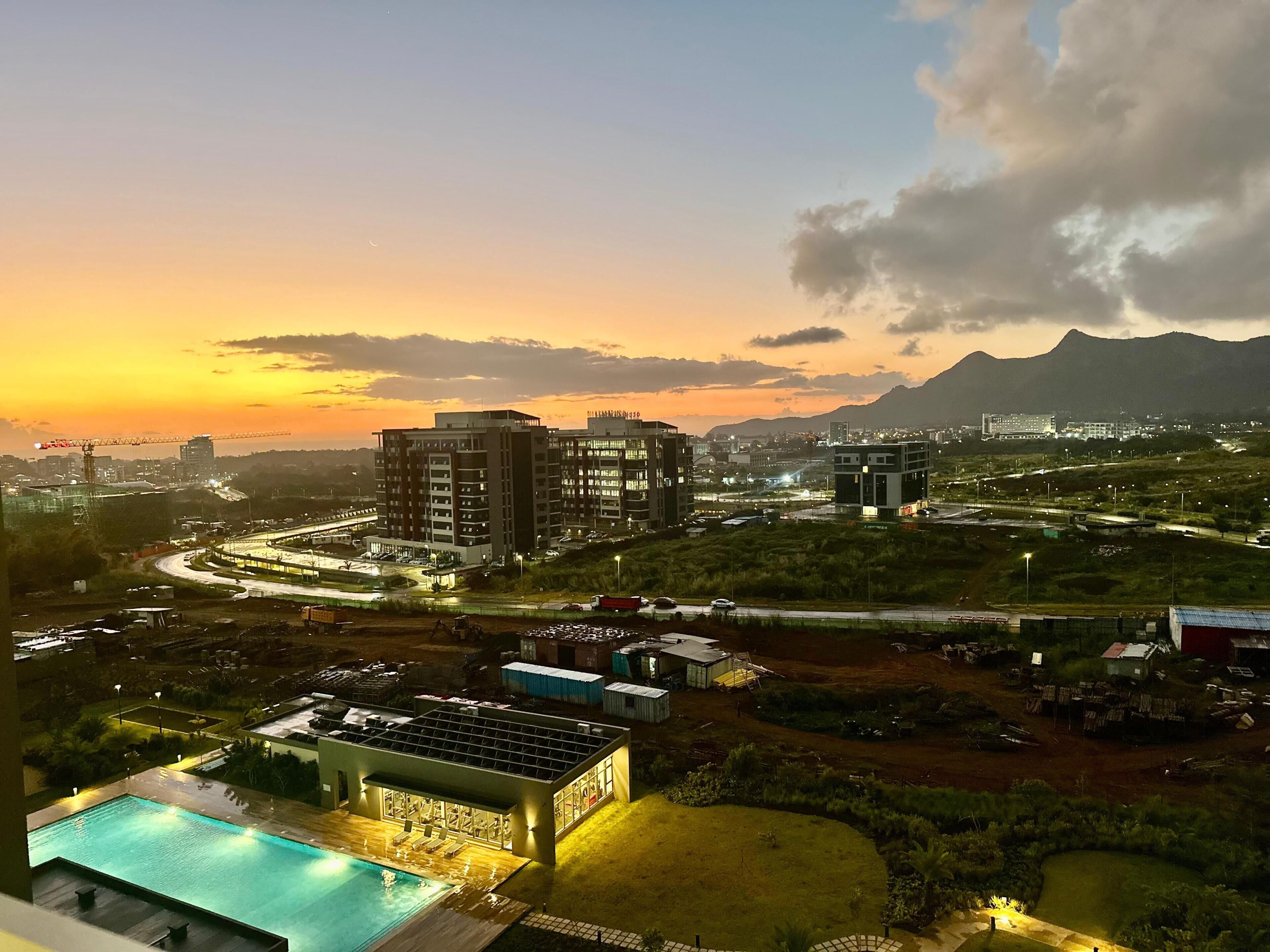 A modern hotel with a swimming pool, a landscaped garden, and a view of the cityscape at sunset.