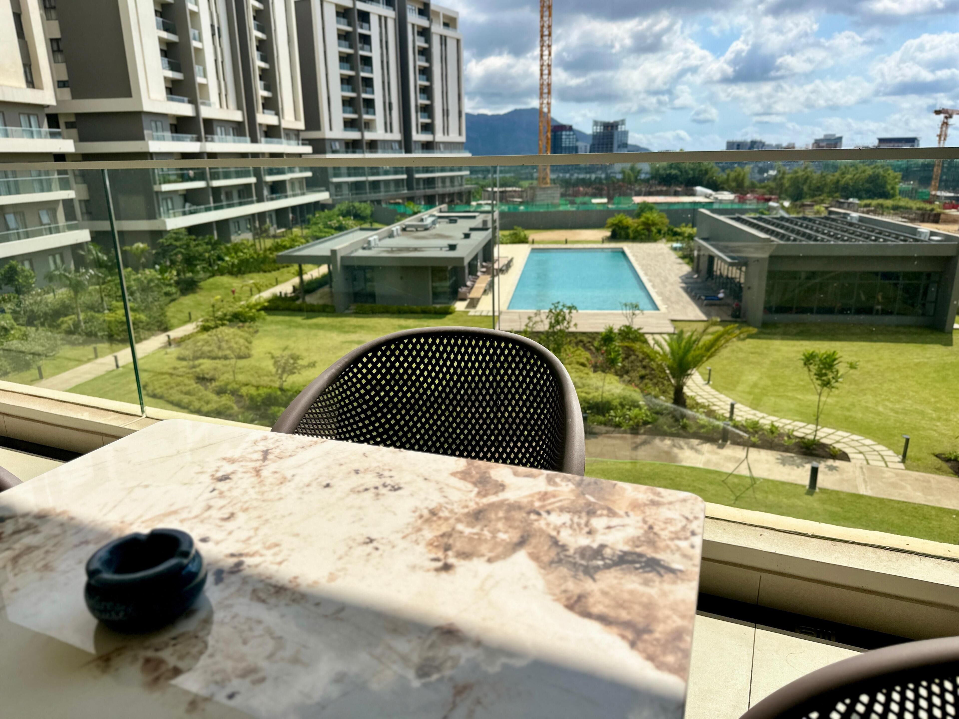 Habitación de hotel moderna con una mesa de mármol, una silla negra y vistas a una piscina, vegetación y otros edificios.