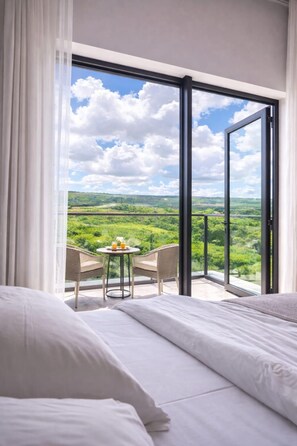 A hotel room with a large sliding glass door leading to a balcony with a table and chairs, offering a view of a green landscape. - Minissy Suites (Moka)