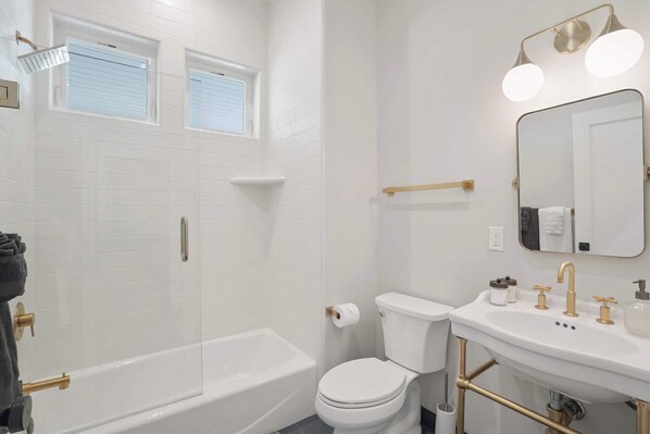 Un baño moderno con ducha de vidrio, inodoro blanco y lavamanos con detalles dorados.
