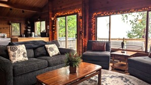 A cozy cabin interior with a wooden ceiling, a sofa, a chair, a coffee table, and a view of the outdoors through large windows. - Waterfront, Rustic, Hot Tub, Pet-Friendly (Sequim)