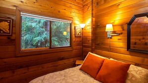 A wooden cabin room with a bed, two wall-mounted lamps, and a window with a view of trees. - Waterfront, Rustic, Hot Tub, Pet-Friendly (Sequim)