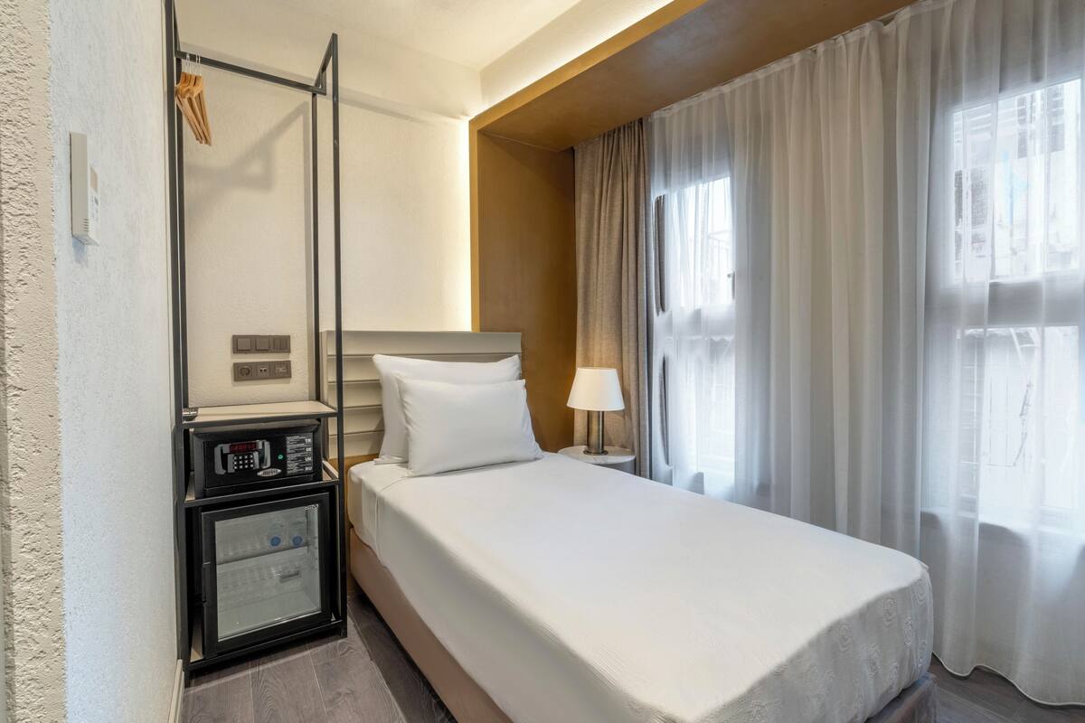 A hotel room with a bed, a nightstand, a mini-fridge, a lamp, and a window with curtains.
