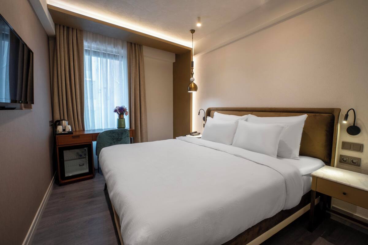 A hotel room with a large bed, a bedside table, a nightstand, a TV mounted on the wall, and a window with curtains.