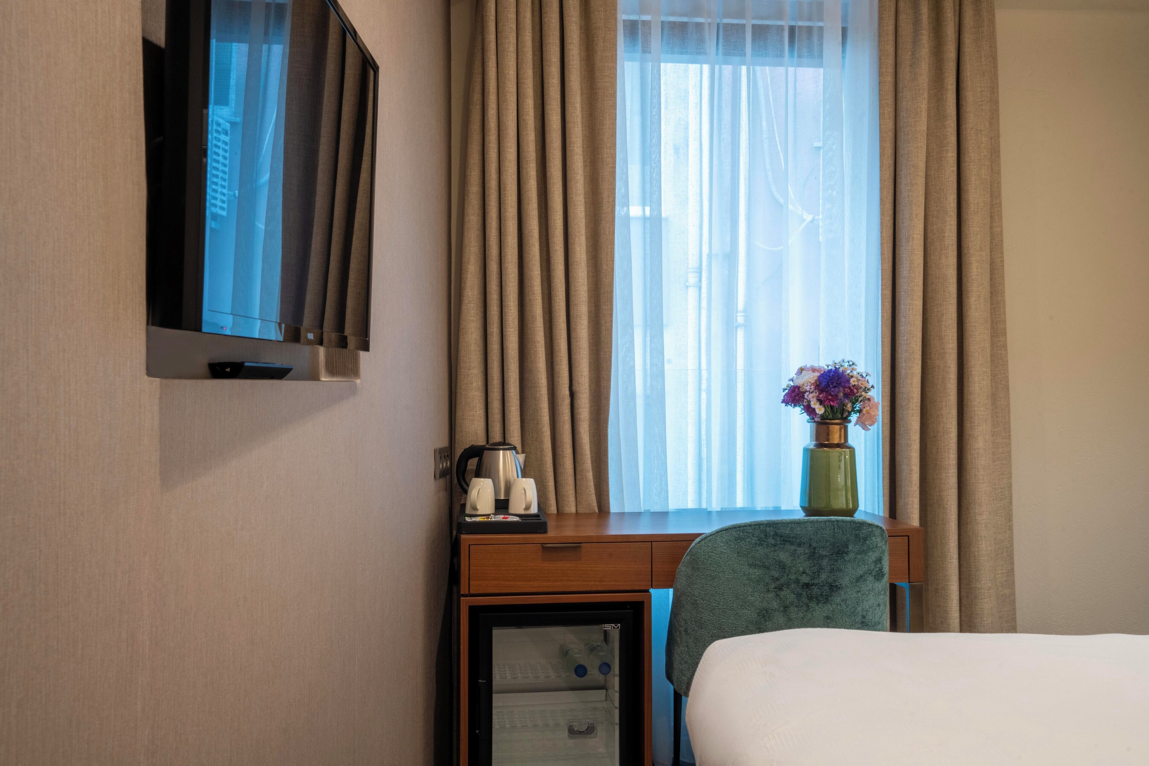 A hotel room with a bed, a desk with a laptop, a coffee maker, and a vase with flowers.
