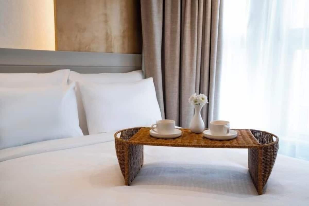 A hotel room with a bed, a tray table with two cups and a vase, and a window with curtains.
