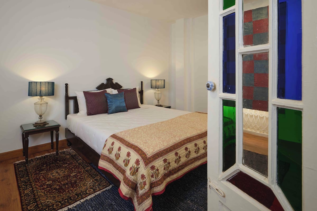 Une chambre à coucher avec un lit, deux tables de chevet avec des lampes, un tapis à motifs et une fenêtre donnant sur un paysage coloré.