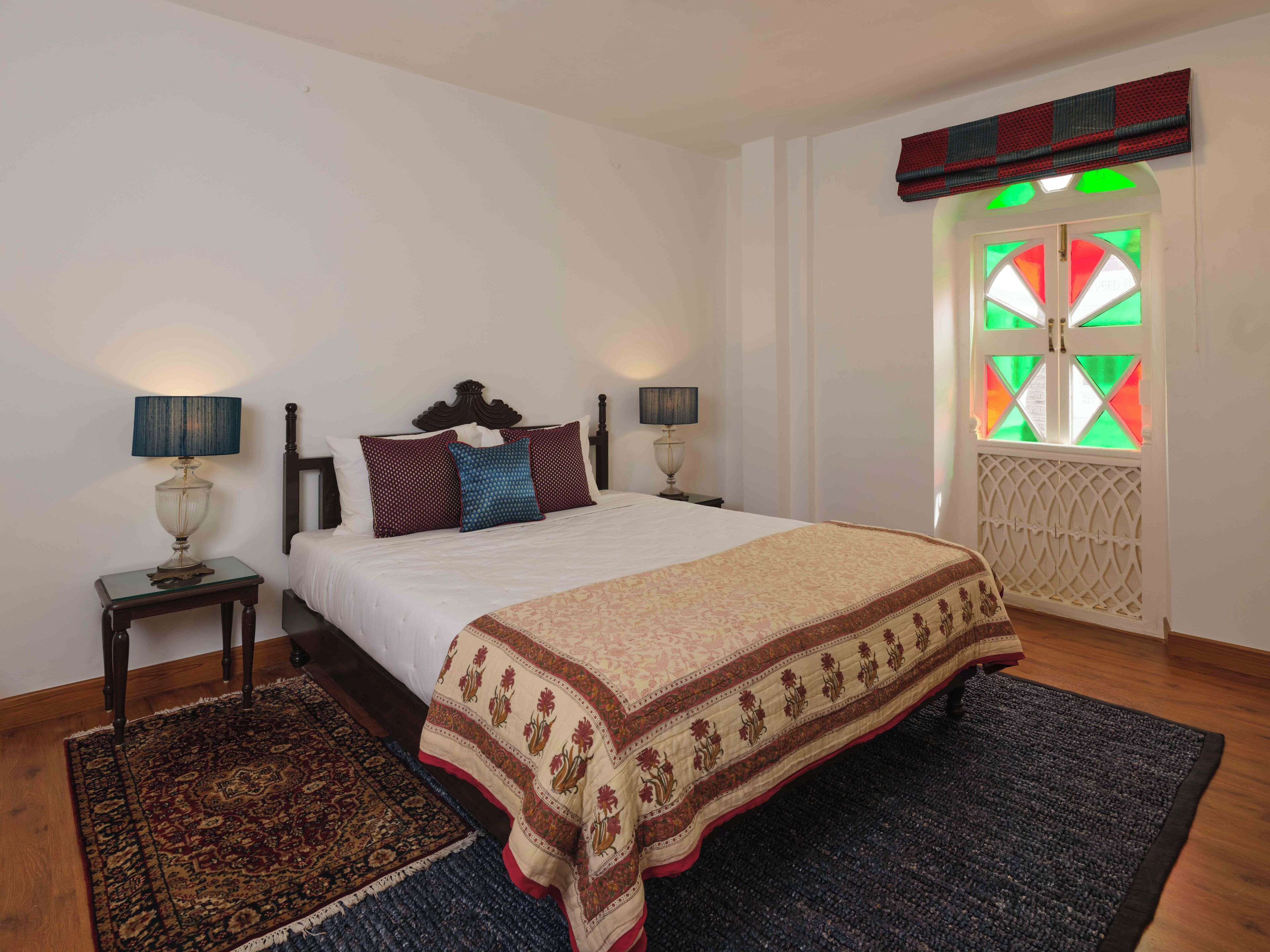 A bedroom with a bed, two bedside tables, a nightstand, a window with a geometric pattern, and a decorative ceiling.