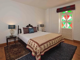 A bedroom with a bed, two bedside tables, a nightstand, a window with a geometric pattern, and a decorative ceiling.