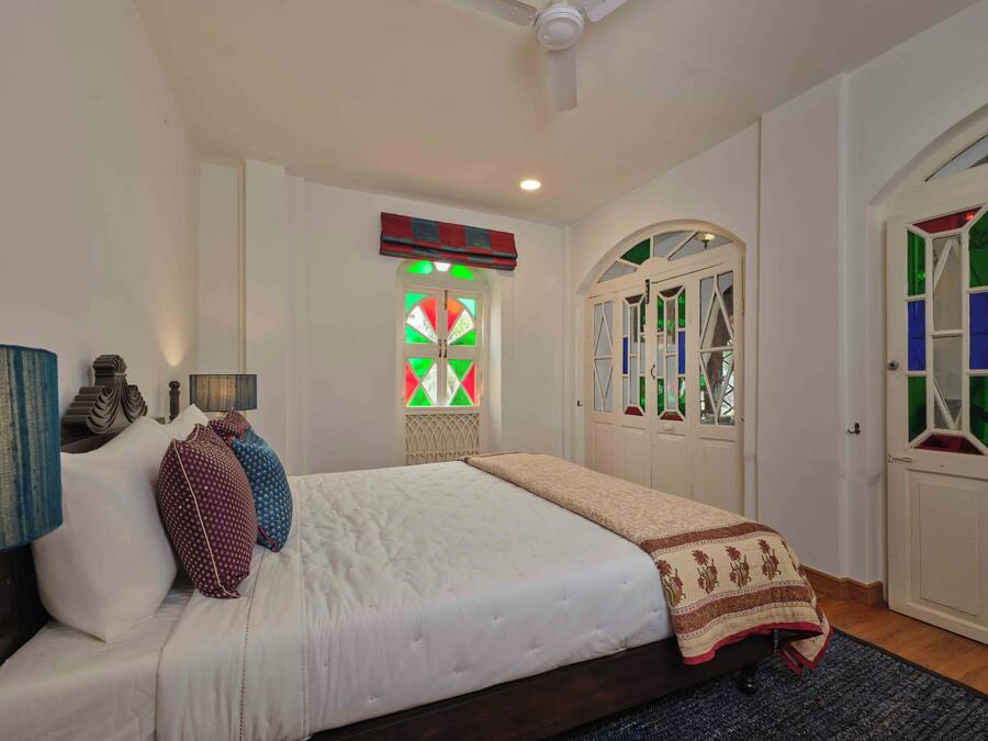 A bedroom with a large bed, a ceiling fan, a window with colorful stained glass, and a wardrobe with glass doors.