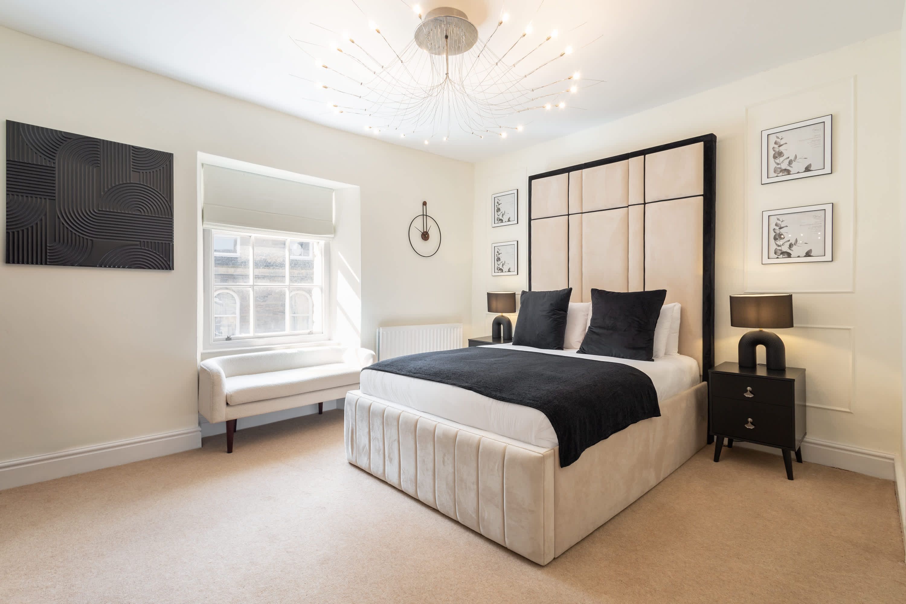 A modern bedroom with a large bed, a bench, a bedside table, a wall clock, and framed pictures.