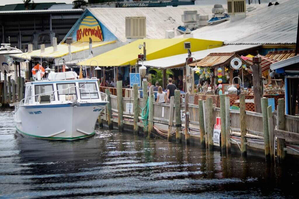 Una zona lungo il fiume con un'imbarcazione ormeggiata, un molo con negozi e un edificio con il cartello 'Riverwalk'.