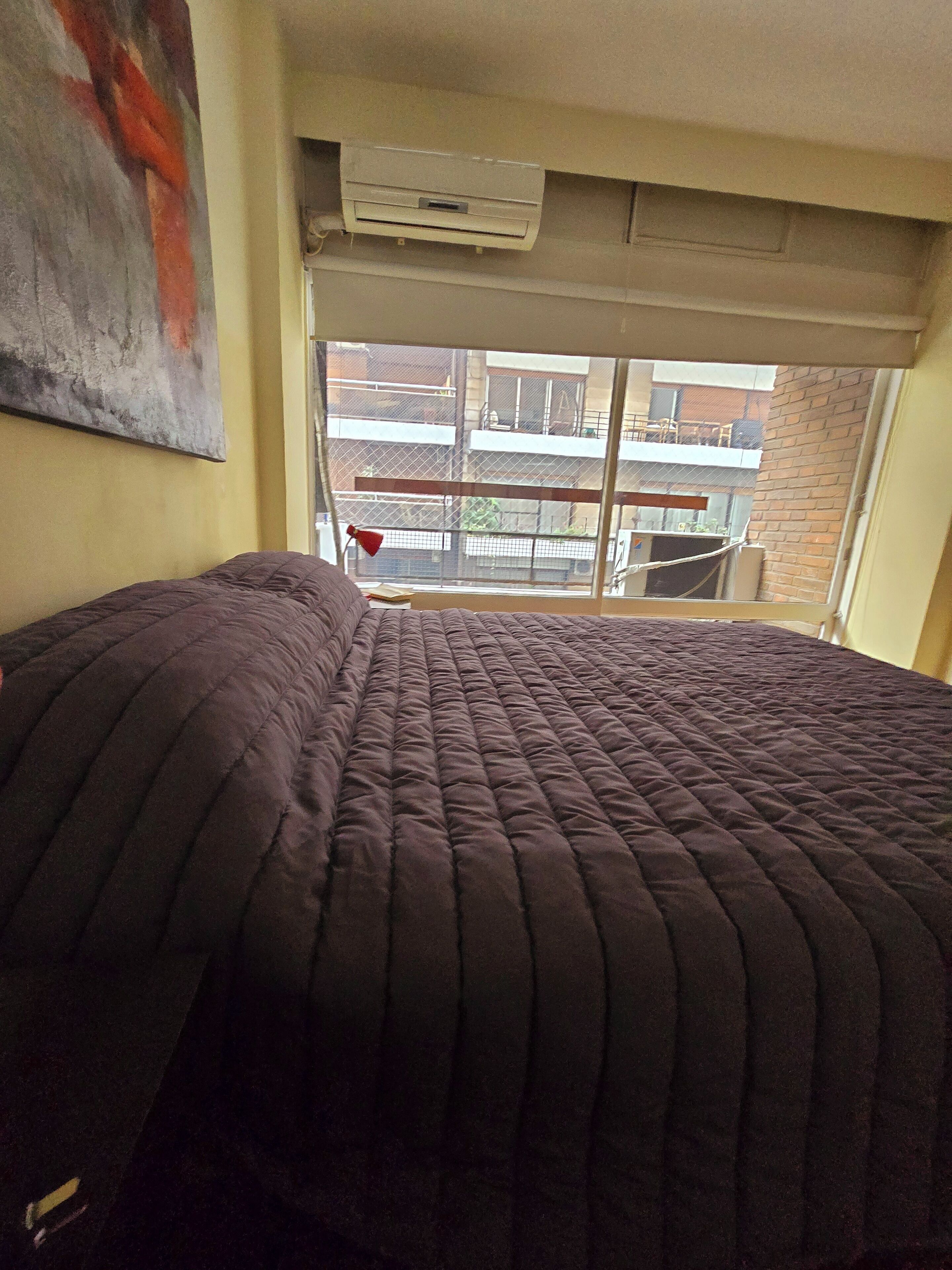 Ein Bett mit einer dunklen Tagesdecke, ein Fenster mit Blick auf einen Balkon und eine Klimaanlage über dem Fenster.