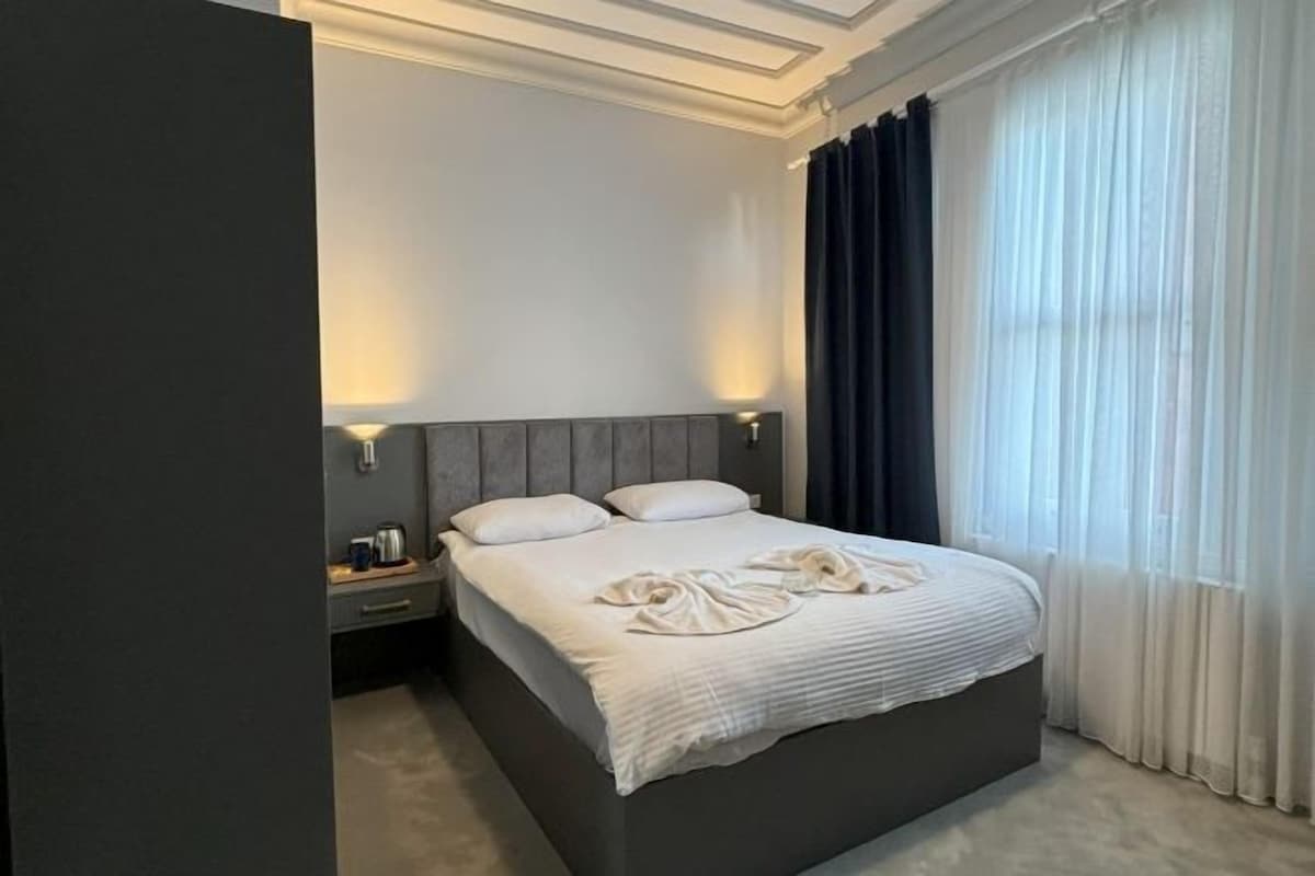 A neatly made bed with a grey headboard, a bedside table with a lamp, and a window with sheer curtains.
