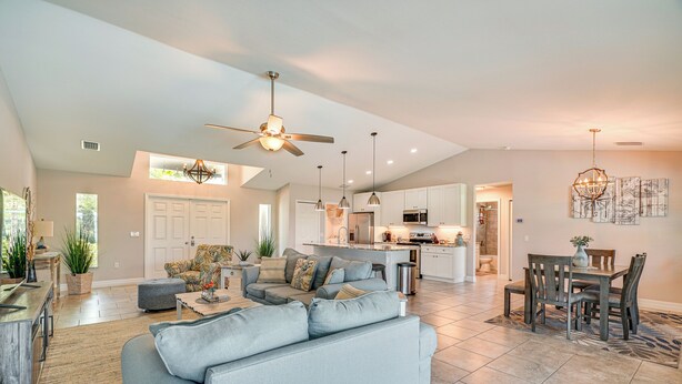 A spacious living area with a grey sofa, dining table, and chairs. There is a kitchen in the background with white cabinets and a central island. - Cape Coral Home w/ Lanai ~ 7 Mi to Marina! (Cape Coral)