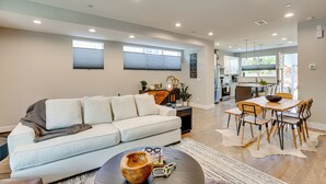 A modern living room with a sofa, coffee table, and dining area with chairs. There is a kitchen in the background with cabinets and a window. - Hot Tub & Mtn-view Rooftop Deck: Golden Home (Golden)