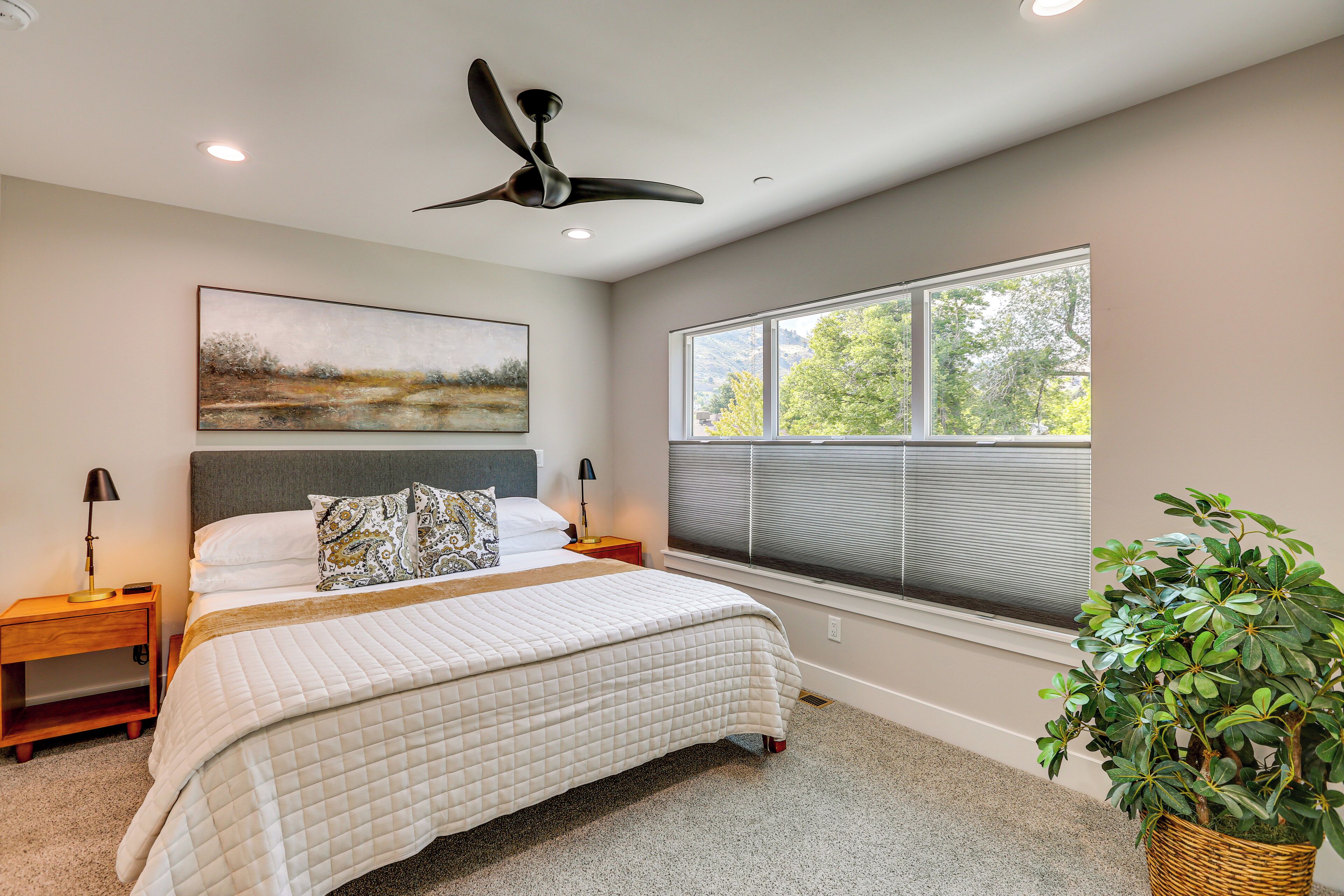 A bedroom with a bed, a ceiling fan, a window with blinds, a nightstand, a lamp, and a potted plant.