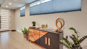 A modern room with a wooden floor, a dark-colored console table with decorative items, a potted plant, and a large window with blinds. - Hot Tub & Mtn-view Rooftop Deck: Golden Home (Golden)
