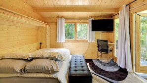 Ein Zimmer in einer Holzhütte mit einem Bett, einem Schreibtisch, einem Sessel, einem Fernseher und einem Fenster mit Vorhängen.
