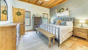 A bedroom with a wooden ceiling, a bed with white bedding, a bench, a dresser, and a nightstand with a lamp. - Ruidoso Retreat w/ Hot Tub & Patio! (Ruidoso)