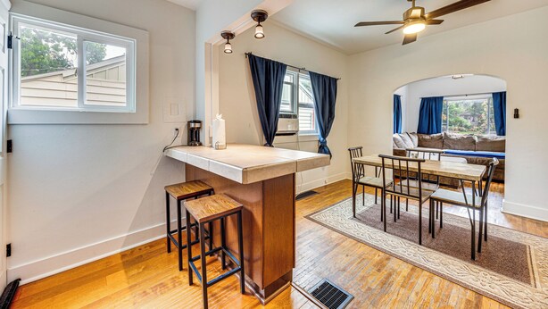 A kitchen with a bar area, wooden stools, a dining table with chairs, and a ceiling fan.