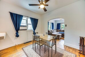 Een eethoek met een houten tafel en stoelen, een plafondventilator, en uitzicht op een woonkamer met een bank en een raam met blauwe gordijnen.