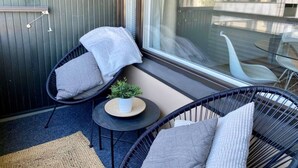 Un balcon avec deux fauteuils en osier noir, une petite table ronde ornée d’une plante, et une vue sur les immeubles à travers une porte vitrée.