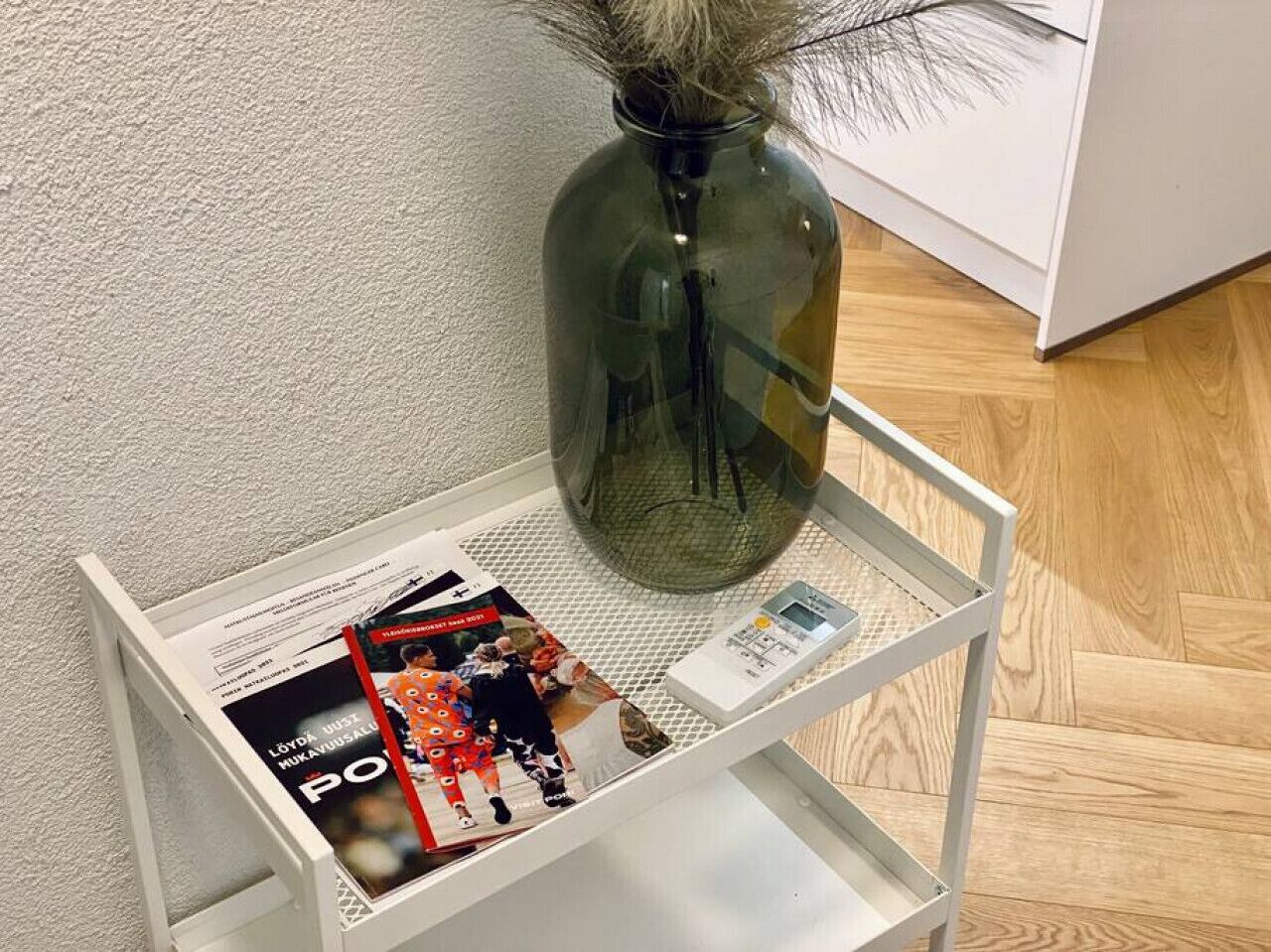 A white metal shelf with a magazine, a small vase with dried plants, and a white object on a textured wall.