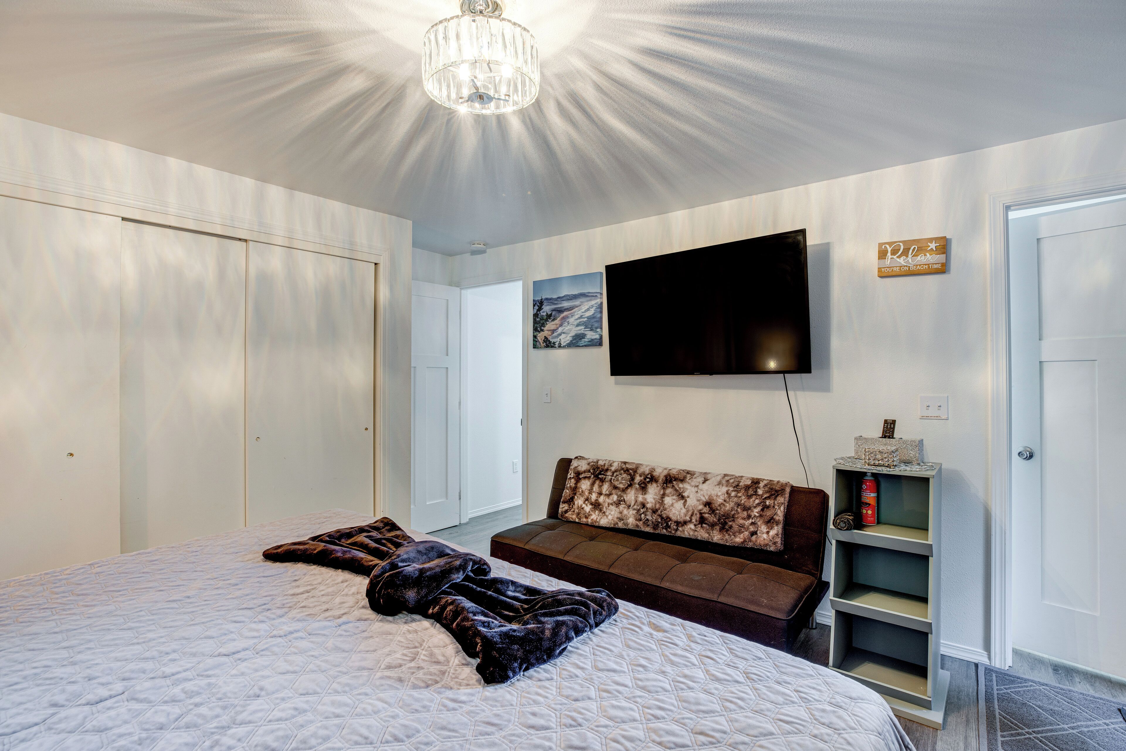 A bedroom with a large bed, a sofa, a TV mounted on the wall, and a small shelf with items.