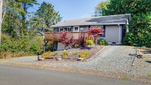 A house with a wooden deck, a gravel driveway, and a small garden with various plants. - Rockaway Beach House - Walk to Ocean! (Rockaway Beach)