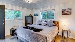 A bedroom with a large bed, two windows, a chandelier, a side table, and a cabinet. - Rockaway Beach House - Walk to Ocean! (Rockaway Beach)