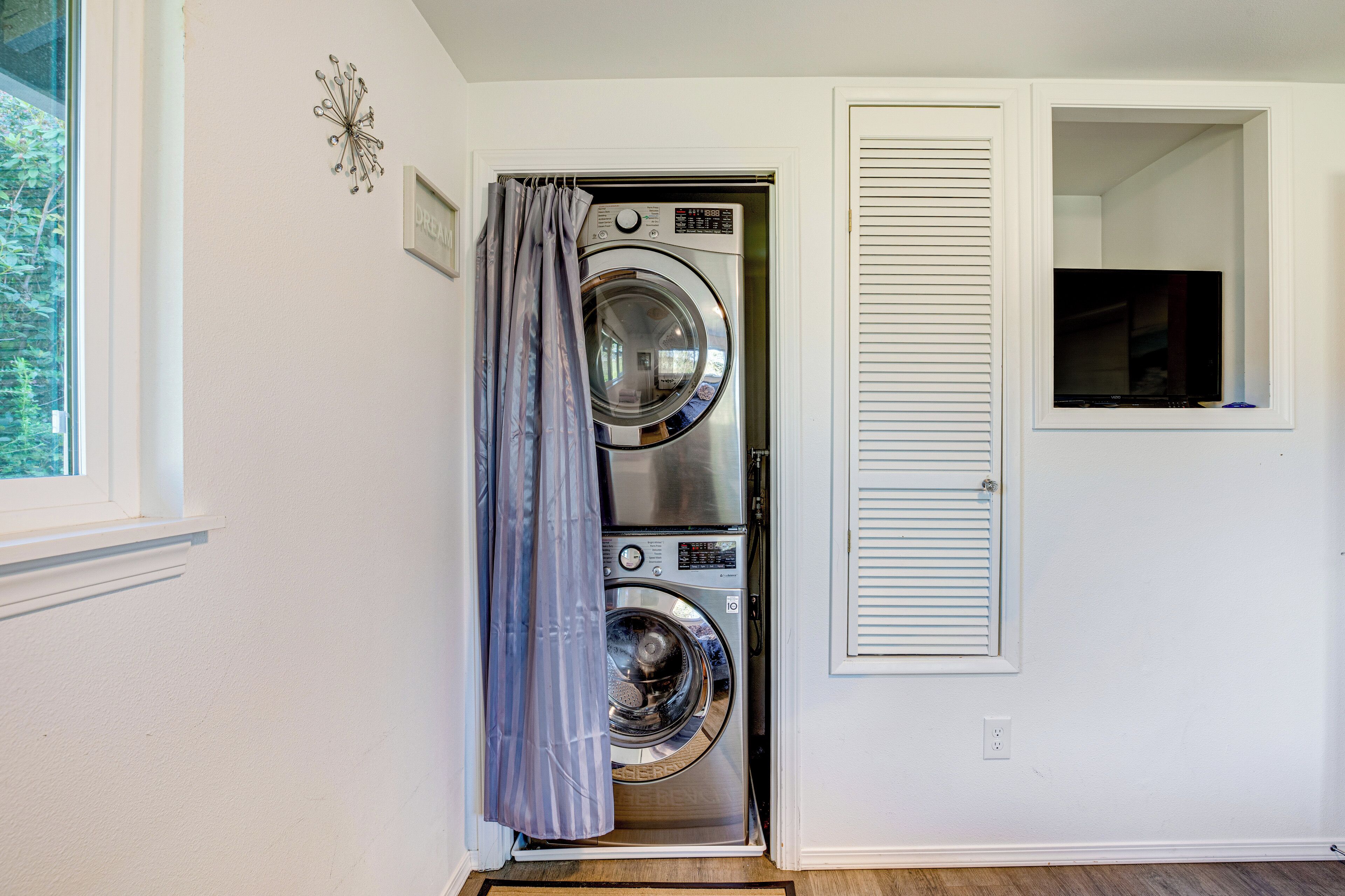 A laundry room with a front-loading washer and dryer, a window with blinds, and a door with a curtain.