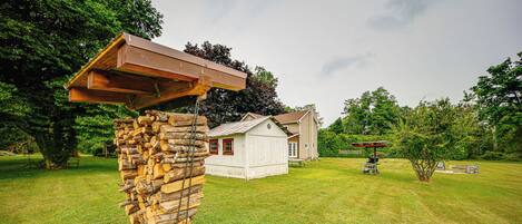 A rustic wooden structure with a slanted roof, a white building with a red door, and a well-maintained lawn in the background.
