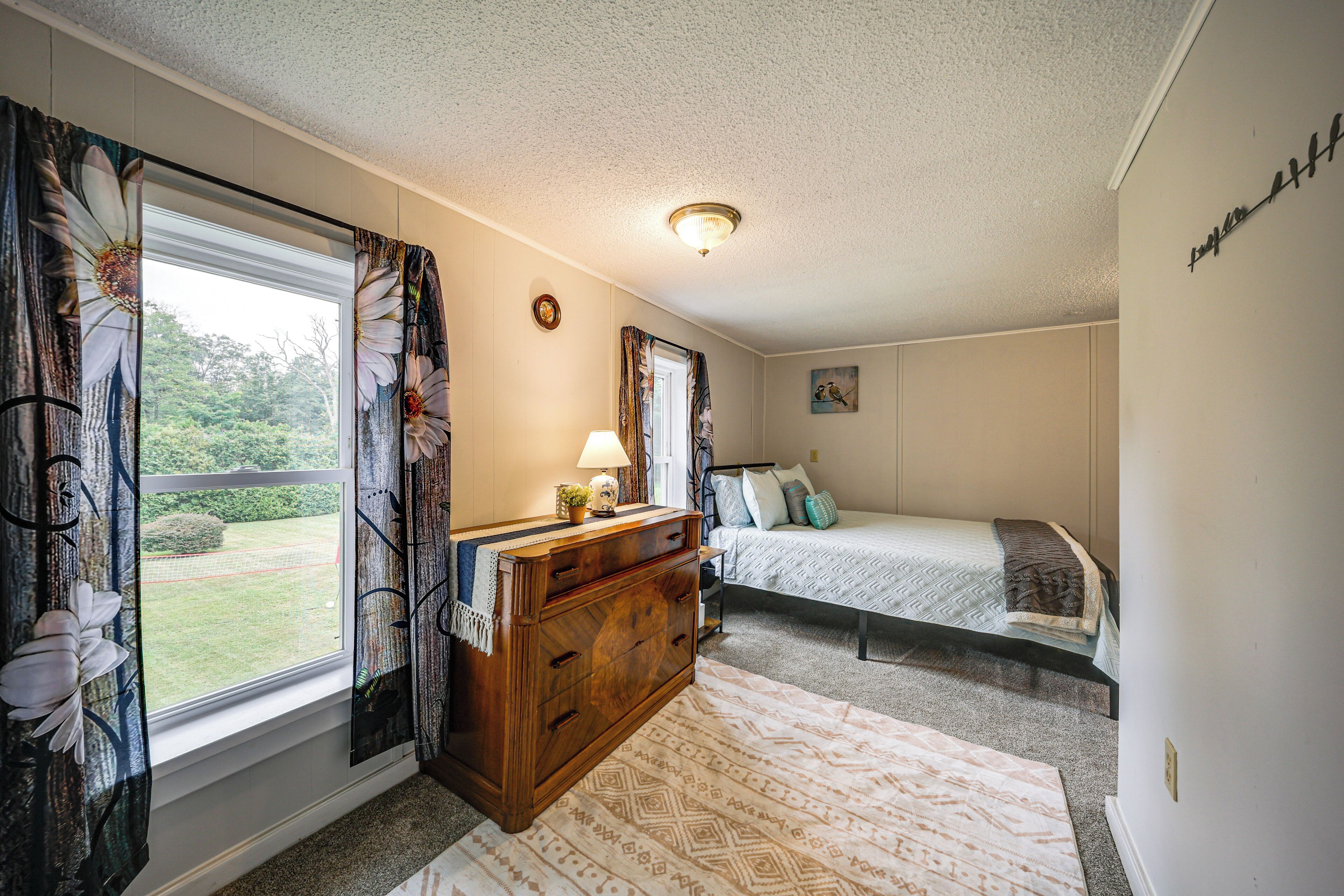 A bedroom with a bed, a dresser, a window with curtains, a lamp, and a painting on the wall.