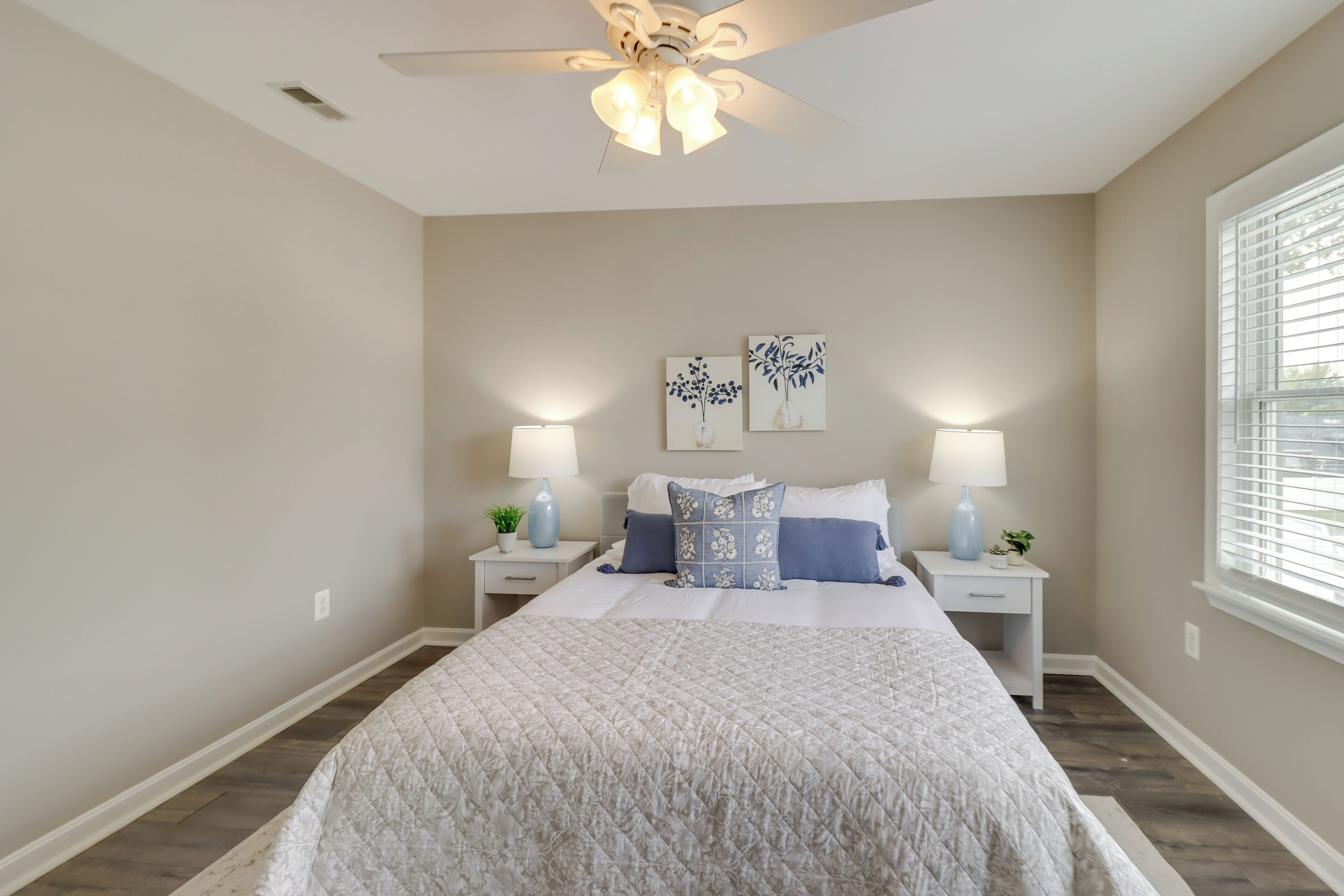 A bedroom with a large bed, two bedside tables with lamps, a ceiling fan, and artwork on the wall.