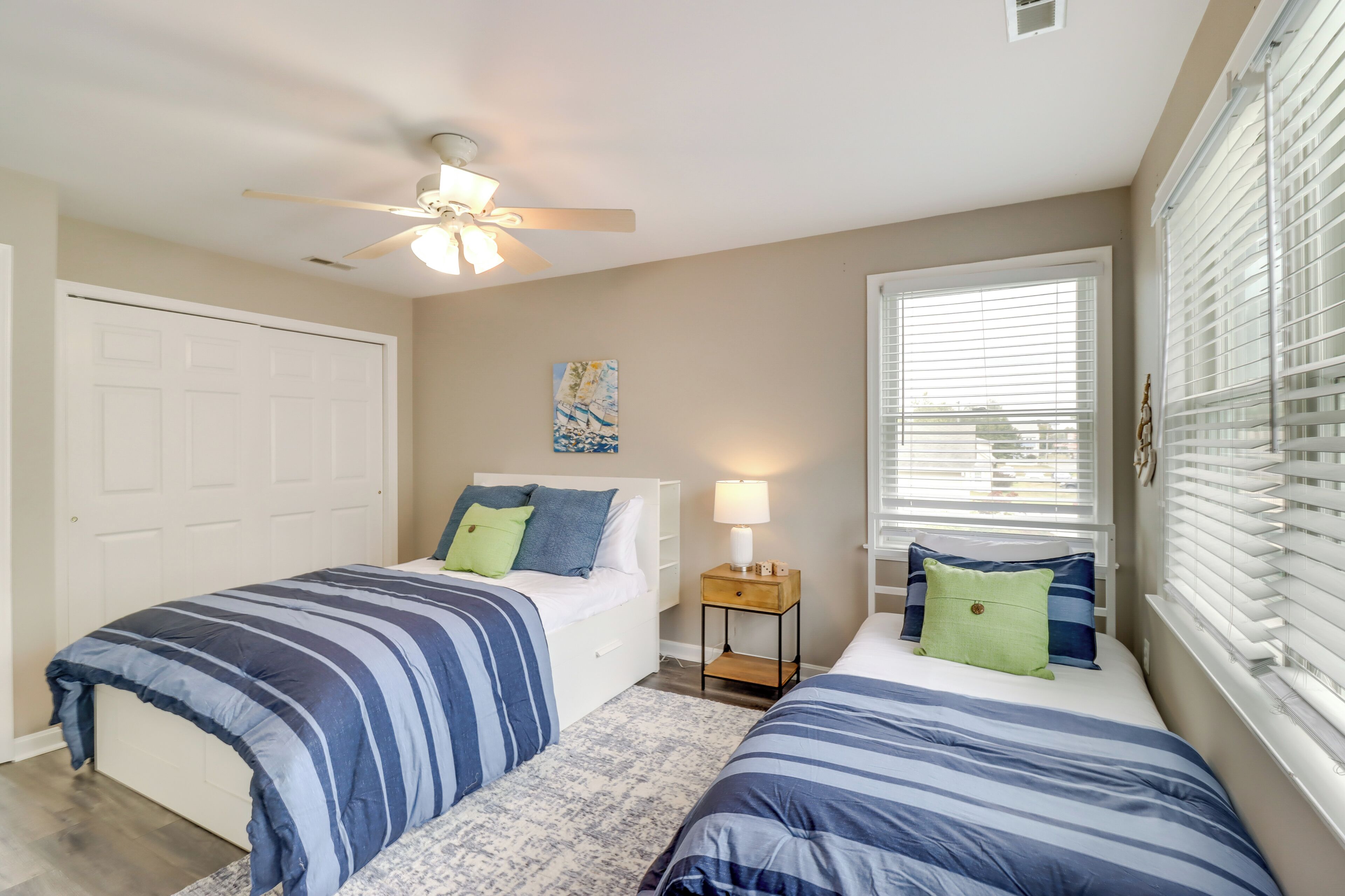 A bedroom with two beds, a ceiling fan, a nightstand with a lamp, and a window with blinds.