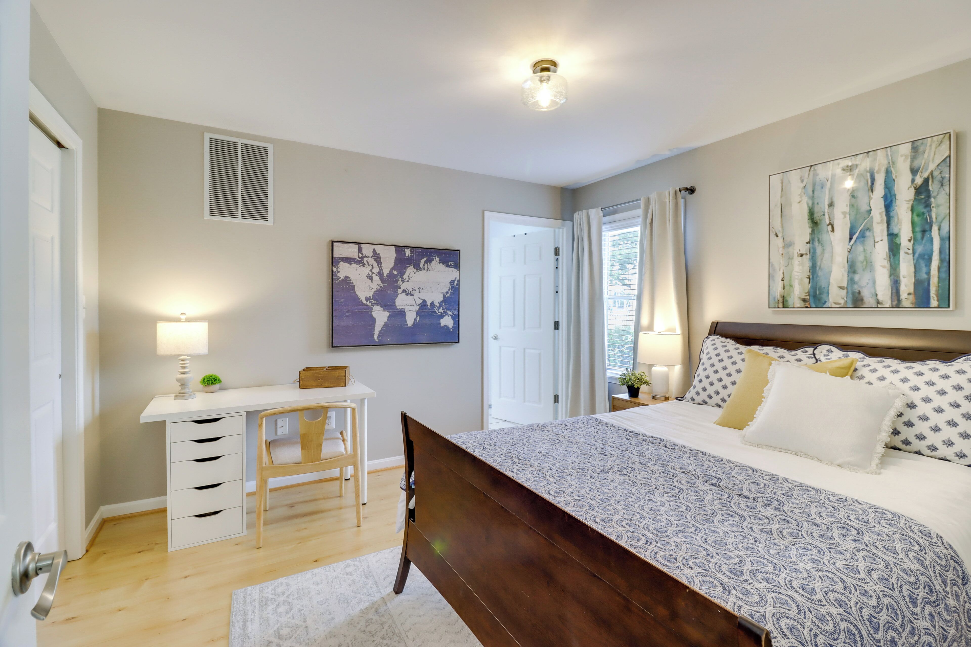 A bedroom with a wooden bed, a desk with a chair, a world map on the wall, and a large window with curtains.
