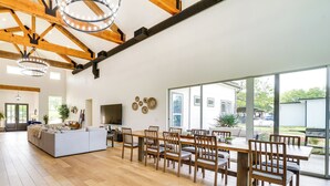 A spacious living area with a large sofa, dining table, and chairs. There is a television and decorative wall art. The room has wooden beams and large windows. - Lakefront Texas Home on Legacy Ranch (D'Hanis)