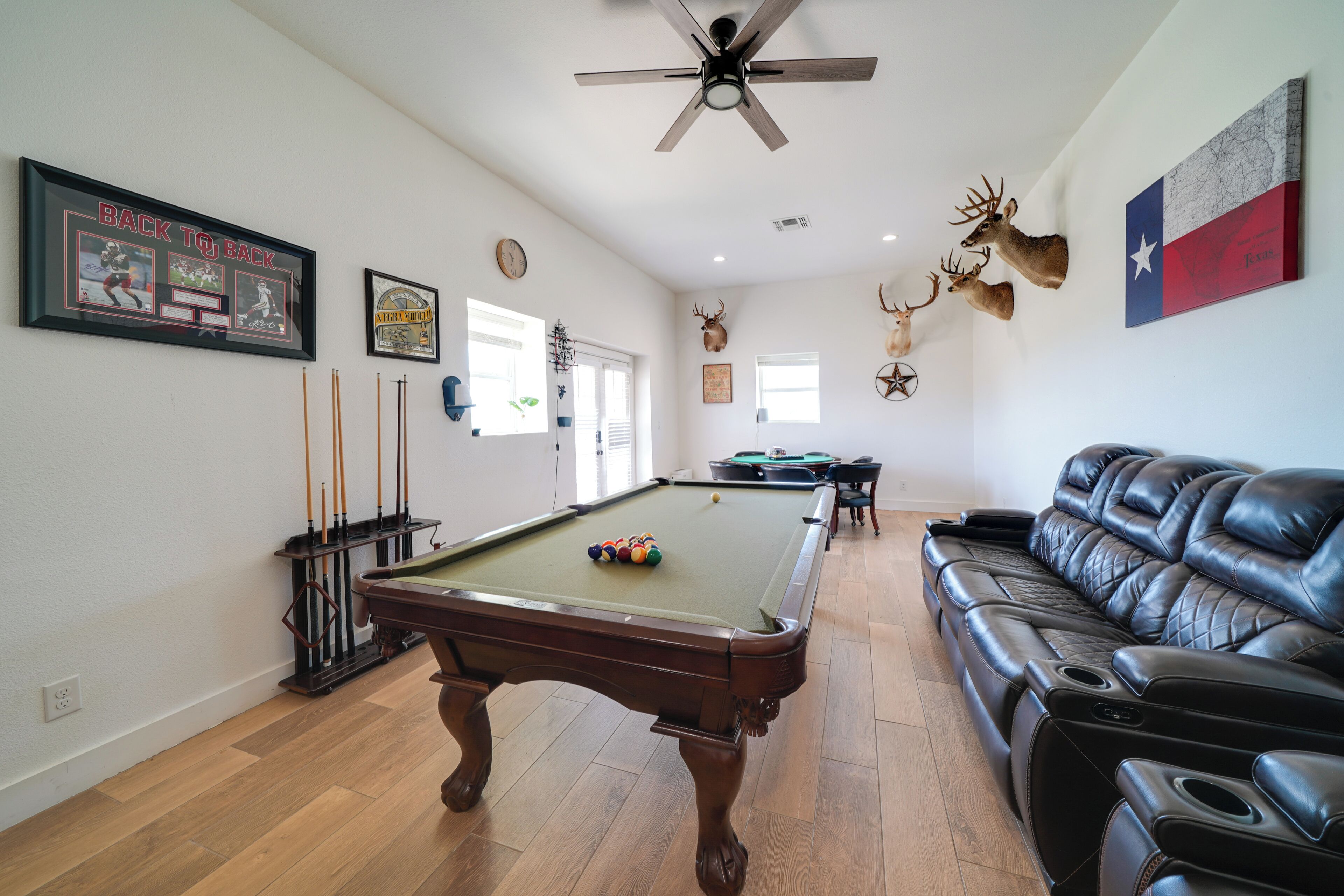 A pool table in a room with a leather couch, a ceiling fan, and wall-mounted deer heads.