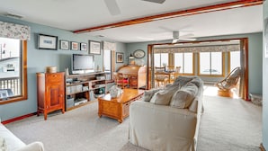 A spacious living room with a large sofa, wooden coffee table, and a television. There are bookshelves, a fireplace, and large windows with curtains. - Lovely Coastal Home By Delaware Bay: Walk to Beach (Fortescue)