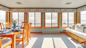 A room with a dining table set for four, a sofa, and large windows overlooking a body of water. - Lovely Coastal Home By Delaware Bay: Walk to Beach (Fortescue)
