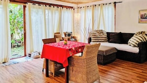 Ein gemütliches Zimmer mit Holzbalkendecke, einem Esstisch mit roter Tischdecke, Korbmöbeln und einer Couch mit gestreiften Kissen.