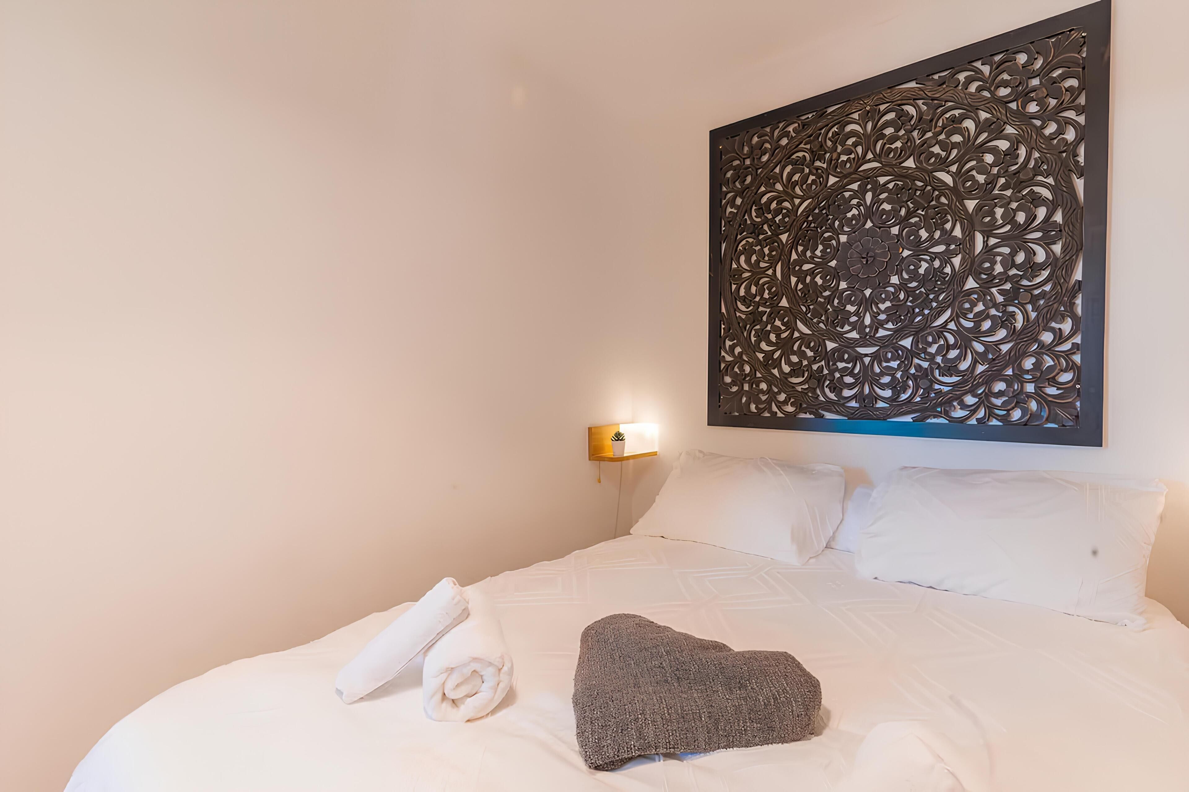 A neatly made bed with white linens and a gray throw blanket, a decorative wall panel, and a wall-mounted light fixture.