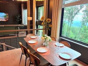 A dining area with a table set for four, a vase with flowers, and a view of greenery outside.