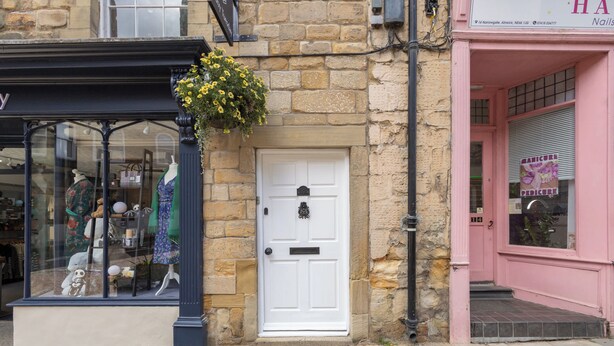 A storefront with a white door, a hanging flower basket, and a pink building across the street. - Host & Stay | Yule House (Alnwick)