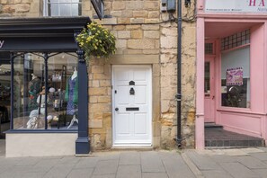 A storefront with a white door, a hanging flower basket, and a pink building across the street. - Host & Stay | Yule House (Alnwick)